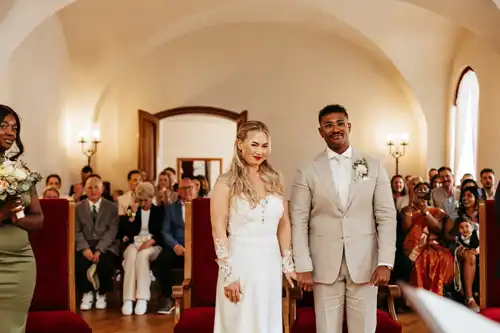 Braut und Bräutigam stehen zusammen am Altar, halten sich an den Händen und lächeln, während die Gäste in einem eleganten, gut beleuchteten Raum mit gewölbten Decken zusehen - wunderschön eingefangen von Hochzeitsfotograf Görlitz