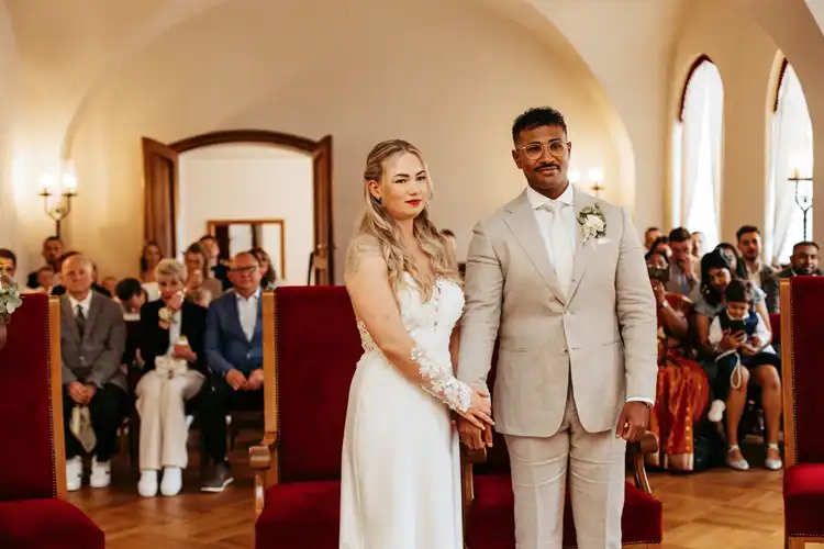 Eine Braut in einem weißen Kleid und ein Bräutigam in einem hellen Anzug stehen händchenhaltend vor einem Raum auf Hochzeit Gut am See, mit sitzenden Gästen im Hintergrund während ihrer Hochzeitszeremonie. Hochzeitsfotograf Görlitz hält diesen besonderen Moment fest.
