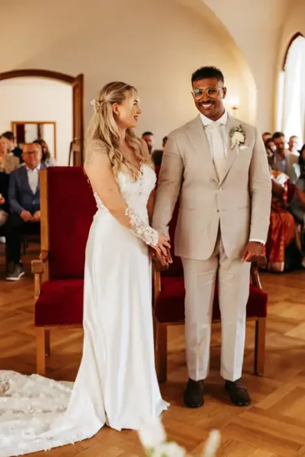 Eine Braut in einem weißen Kleid und ein Bräutigam in einem beigen Anzug halten sich bei ihrer Hochzeit auf Gut am See an den Händen und lächeln sich an, während die Gäste den romantischen Moment beobachten.