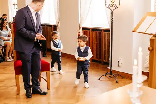 Zwei Jungen in passenden marineblauen Anzügen und weißen Turnschuhen schreiten bei einer Hochzeit auf Gut am See zum Altar, während ein Mann applaudiert und die Gäste in dem hellen Raum zusehen - wunderschön eingefangen von Hochzeitsfotograf Görlitz.