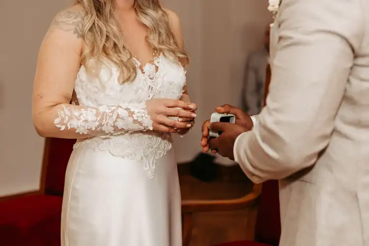 Eine Braut in einem weißen Spitzenhochzeitskleid steht ihrem Bräutigam gegenüber, der während ihrer Hochzeit Gut am See eine offene Ringschachtel hält. Die Gesichter der Braut und des Bräutigams bleiben außerhalb des Bildausschnitts, was diesen intimen Moment unterstreicht.
