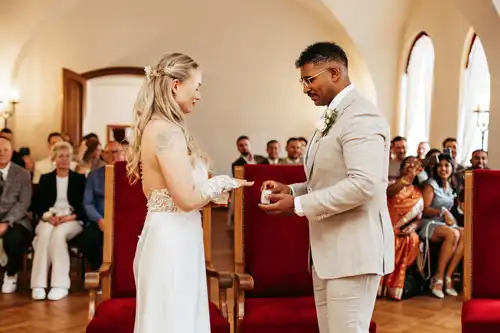 Eine Braut und ein Bräutigam stehen sich gegenüber und tauschen bei ihrer Hochzeit in Görlitz in einem hellen Raum mit sitzenden Gästen, die lächelnd zusehen, die Ringe aus.
