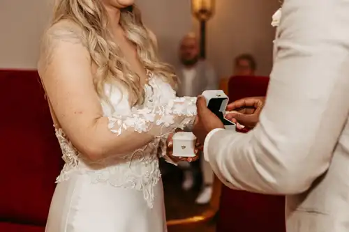 Eine Braut in einem weißen Spitzenhochzeitskleid und ein Bräutigam in einem hellen Anzug tauschen während ihrer Hochzeitszeremonie auf Gut am See die Ringe aus, wobei jeder eine kleine Ringschachtel in der Hand hält. Schön eingefangen von Hochzeitsfotograf Görlitz.
