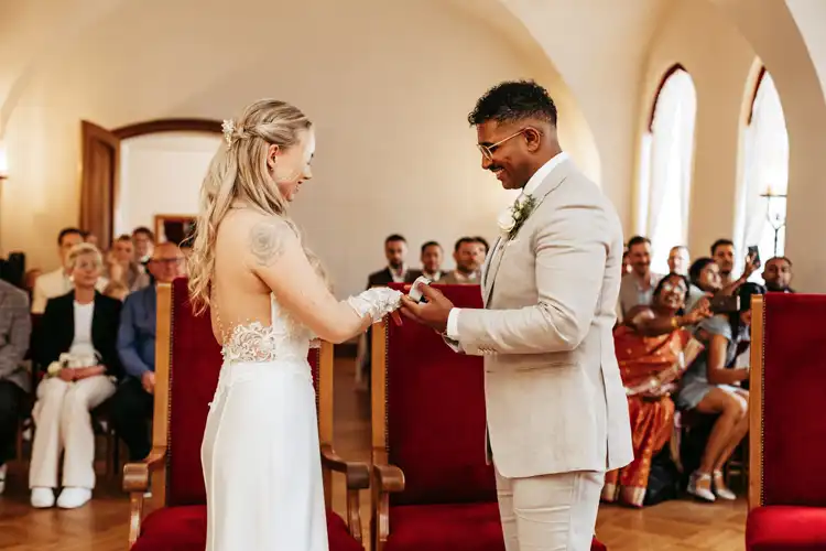 Eine Braut und ein Bräutigam tauschen während einer Hochzeit Gut am See in einem hellen Raum die Ringe aus und lächeln sich dabei an. Die Gäste sitzen im Hintergrund und beobachten den besonderen Moment des Paares, der von Hochzeitsfotograf Görlitz festgehalten wurde.