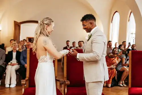 Eine Braut und ein Bräutigam tauschen während einer Hochzeit Gut am See in einem hellen Raum die Ringe aus und lächeln sich dabei an. Die Gäste sitzen im Hintergrund und beobachten den besonderen Moment des Paares, der von Hochzeitsfotograf Görlitz festgehalten wurde.