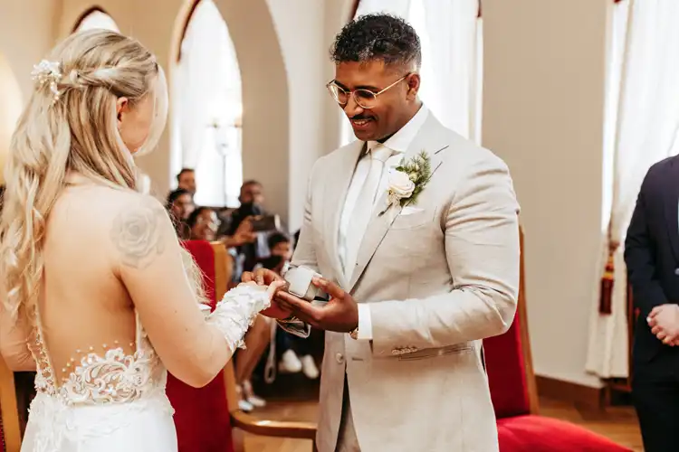 Ein Bräutigam in einem hellen Anzug steckt seiner Braut während einer Hochzeit auf Gut am See einen Ring an den Finger. Die Braut trägt ein rückenfreies weißes Kleid mit Spitzendetails, während im Hintergrund Gäste und Ihr Hochzeitsfotograf Görlitz sitzen.