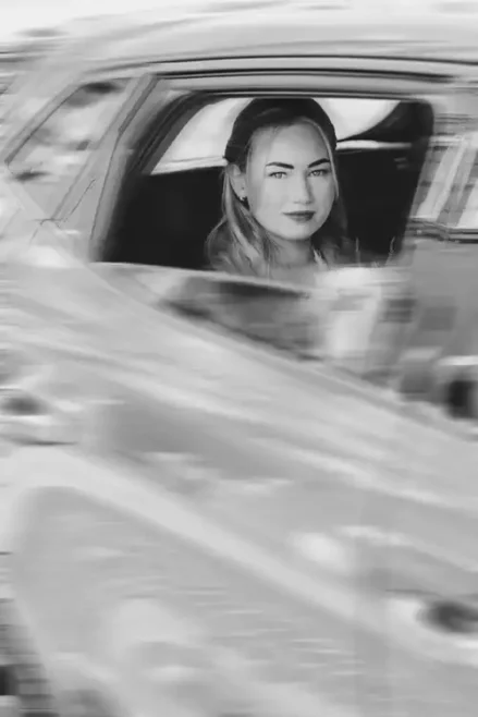 Eine Frau mit langen Haaren sitzt auf dem Rücksitz eines Autos und schaut mit einem leichten Lächeln aus dem Fenster. Das von Hochzeitsfotograf Görlitz aufgenommene Schwarz-Weiß-Bild zeigt Spiegelungen in der Autotür und erinnert an zeitlose Momente .
