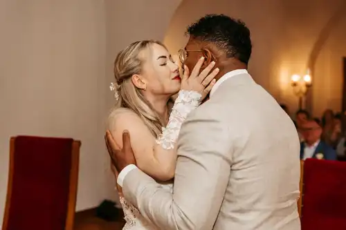 Eine Braut und ein Bräutigam küssen sich während ihrer Hochzeit auf Gut am See und halten sich gegenseitig fest. Die Braut hat helles Haar und trägt ein weißes Kleid, der Bräutigam trägt einen hellen Anzug. Im Hintergrund sind sitzende Gäste zu sehen.