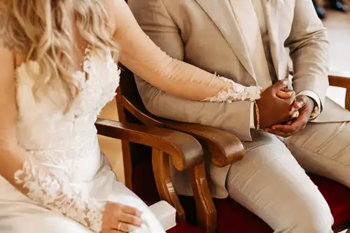 Braut und Bräutigam sitzen nebeneinander auf Stühlen und halten sich bei ihrer Hochzeit auf Gut am See an den Händen. Die Braut trägt ein weißes Spitzenhochzeitskleid, der Bräutigam einen beigen Anzug. Aufgenommen von Hochzeitsfotograf Görlitz, sind die Gesichter der beiden auf dem Bild nicht zu sehen.