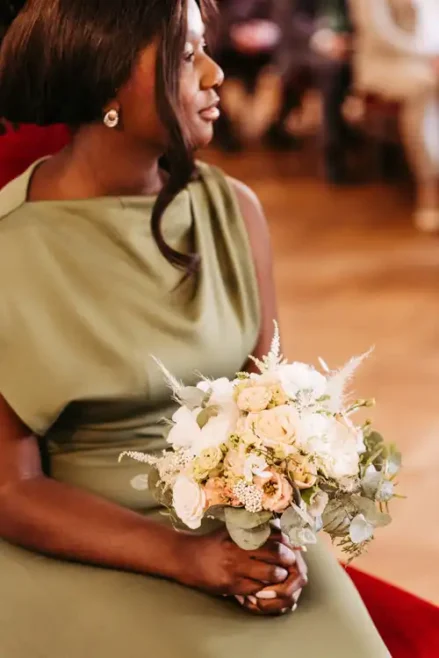 Eine Frau in einem salbeigrünen Kleid hält einen Strauß weißer und blassrosa Blumen mit Grünzeug bei einer Hochzeit auf Gut am See, in einem Innenraum mit unscharfen Menschen im Hintergrund.