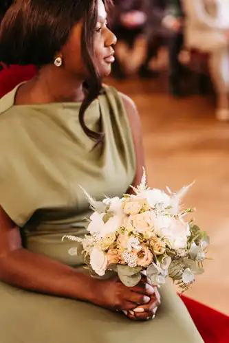 Eine Frau in einem salbeigrünen Kleid hält einen Strauß weißer und blassrosa Blumen mit Grünzeug bei einer Hochzeit auf Gut am See, in einem Innenraum mit unscharfen Menschen im Hintergrund.