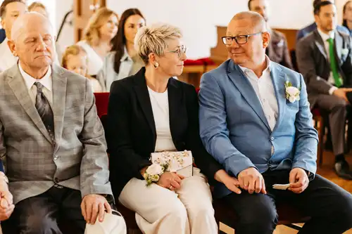 Auf Hochzeit im Standesamt Görlitz sitzen die Gäste in Reihen. Zwei Personen im Vordergrund, aufgenommen von Hochzeitsfotograf Görlitz, lächeln sich herzlich an, während die Gäste im Hintergrund aufmerksam zuhören.