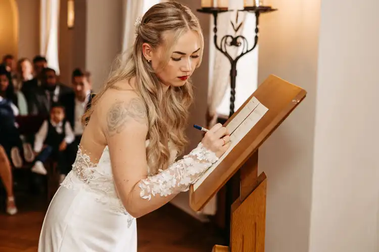Eine Braut mit langen blonden Haaren und einem Spitzenhochzeitskleid unterschreibt eine Urkunde auf einem Holzständer während ihrer Hochzeit auf Gut am See, mit sitzenden Gästen im Hintergrund. Aufgenommen von Hochzeitsfotograf Görlitz.