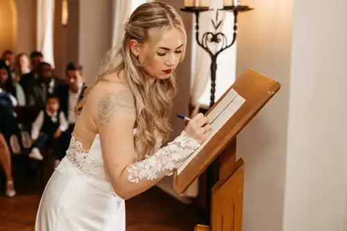Eine Braut mit langen blonden Haaren und einem Spitzenhochzeitskleid unterschreibt eine Urkunde auf einem Holzständer während ihrer Hochzeit auf Gut am See, mit sitzenden Gästen im Hintergrund. Aufgenommen von Hochzeitsfotograf Görlitz.
