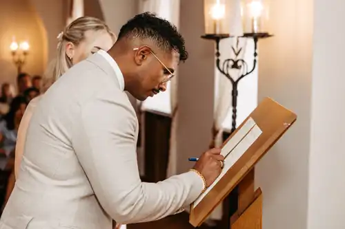 Auf einem Hochzeitsgut am See signiert ein Mann im hellen Anzug ein Dokument auf einem Holzpult, während eine Frau neben ihm steht und das Publikum sowie elegante Kandelaber den Moment beleuchten - festgehalten vom Hochzeitsfotograf Görlitz.