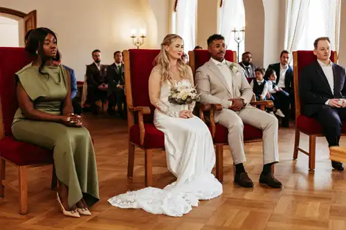 Eine Braut in einem weißen Kleid und ein Bräutigam in einem beigen Anzug sitzen zusammen und halten einen Blumenstrauß, umgeben von Gästen in formeller Kleidung auf der Hochzeit Gut am See, mit roten Stühlen und Bogenfenstern. Aufgenommen von Hochzeitsfotograf Görlitz.