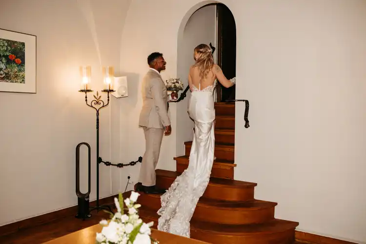 Eine Braut in einem langen weißen Kleid und ein Bräutigam in einem hellen Anzug schreiten händchenhaltend eine geschwungene Holztreppe hinauf, in einem warm beleuchteten Raum mit einem Blumengemälde und kerzenähnlichen Wandleuchten aus dem Standesamt Görlitz.