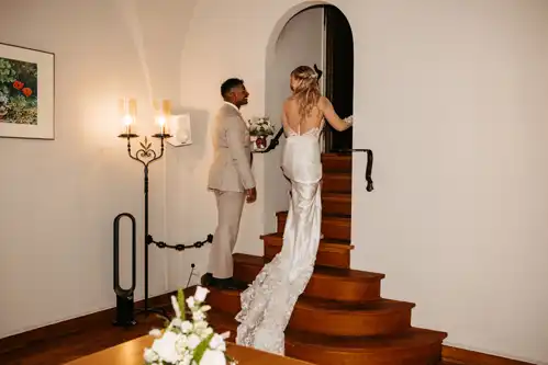 Eine Braut in einem langen weißen Kleid und ein Bräutigam in einem hellen Anzug schreiten händchenhaltend eine geschwungene Holztreppe hinauf, in einem warm beleuchteten Raum mit einem Blumengemälde und kerzenähnlichen Wandleuchten aus dem Standesamt Görlitz.