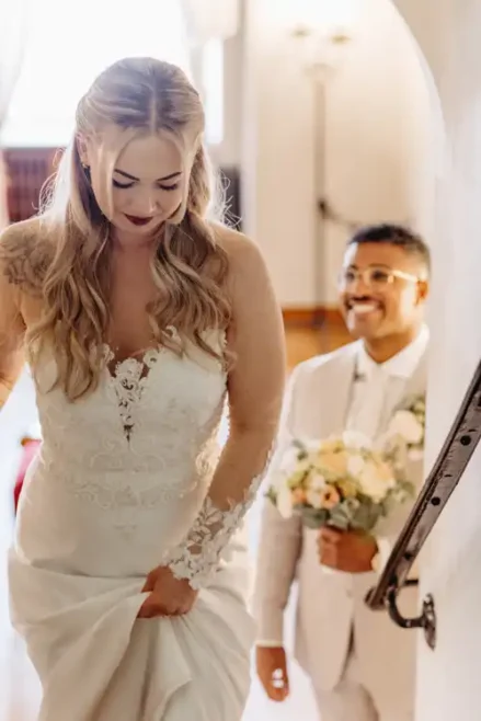 Eine Braut im weißen Spitzenhochzeitskleid geht lächelnd die Treppe hinauf, während der Bräutigam im weißen Anzug und mit Brille mit einem Blumenstrauß hinter ihr herläuft - aufgenommen von Hochzeitsfotograf Görlitz.