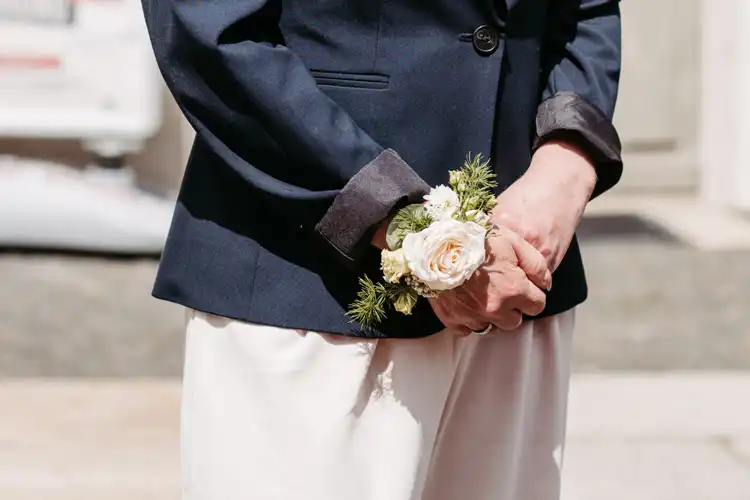 Eine Person in einem marineblauen Blazer und einer cremefarbenen Hose steht mit gefalteten Händen da und hält einen kleinen Strauß weißer und hellgelber Blumen in der Hand - wie geschaffen für eine Hochzeit in Görlitz