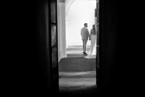 Ein Schwarz-Weiß-Foto zeigt ein händchenhaltendes Paar, das in hellem Licht hinter einer Tür im Standesamt Görlitz steht, eingerahmt von Schatten im Vordergrund, die eine intime, romantische Szene schaffen.