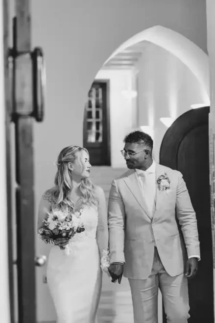 Eine Braut und ein Bräutigam, beide lächelnd, gehen Hand in Hand im Standesamt Görlitz. Die Braut trägt ein langes weißes Kleid und einen Blumenstrauß; der Bräutigam, mit einer Boutonniere, schaut ihr freudig zu - aufgenommen von Hochzeitsfotograf Görlitz.