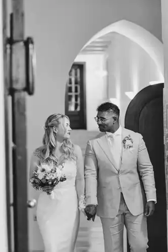 Eine Braut und ein Bräutigam, beide lächelnd, gehen Hand in Hand im Standesamt Görlitz. Die Braut trägt ein langes weißes Kleid und einen Blumenstrauß; der Bräutigam, mit einer Boutonniere, schaut ihr freudig zu - aufgenommen von Hochzeitsfotograf Görlitz.