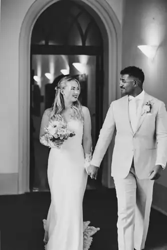Eine Braut und ein Bräutigam, beide lächelnd, gehen Hand in Hand im Standesamt Görlitz. Die Braut trägt ein langes weißes Kleid und einen Blumenstrauß; der Bräutigam, mit einer Boutonniere, schaut ihr freudig zu - aufgenommen von Hochzeitsfotograf Görlitz.