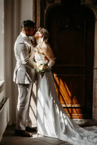 Eine Braut und ein Bräutigam in Hochzeitskleidung küssen sich in einem sonnendurchfluteten Raum auf Hochzeit Gut am See. Die Braut hält einen Blumenstrauß in der Hand und trägt ein langes weißes Kleid mit Schleppe, während der Bräutigam einen hellen Anzug trägt. Hochzeitsfotograf Görlitz hält den Moment fest.