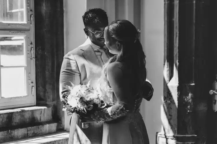 Ein Schwarz-Weiß-Foto eines Paares, das an einem Fenster der Hochzeit Gut am See steht. Die Person auf der linken Seite trägt einen Anzug und eine Brille, während die andere Person in einem Kleid einen Blumenstrauß hält. Aufgenommen von Hochzeitsfotograf Görlitz, wie sie sich gegenseitig anschauen.