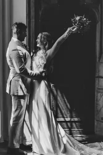 Eine Braut und ein Bräutigam stehen eng beieinander vor einer Holztür auf Hochzeit in Görlitz. Das warme Sonnenlicht, das durch ein Fenster fällt, wirft dramatische Schatten auf den Boden und ihre Hochzeitskleidung.