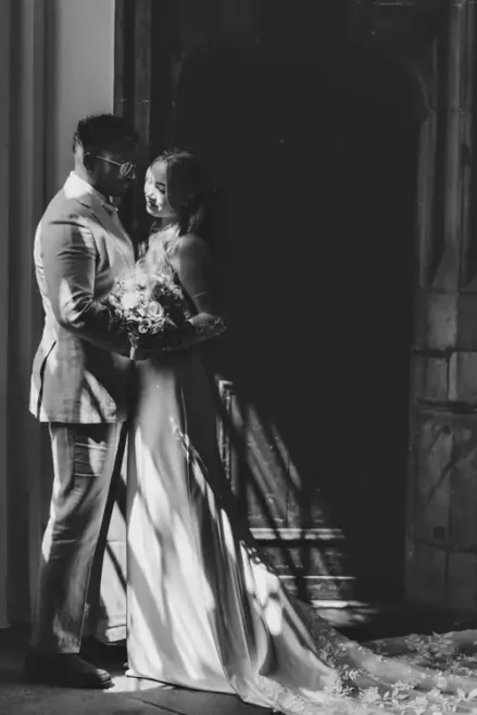 Eine Braut und ein Bräutigam stehen eng beieinander vor einer Holztür auf Hochzeit in Görlitz. Das warme Sonnenlicht, das durch ein Fenster fällt, wirft dramatische Schatten auf den Boden und ihre Hochzeitskleidung.