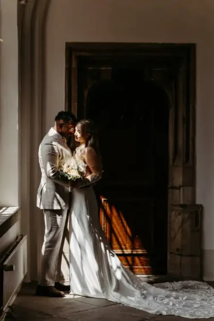 Eine Braut und ein Bräutigam stehen eng beieinander vor einer Holztür auf Hochzeit in Görlitz. Das warme Sonnenlicht, das durch ein Fenster fällt, wirft dramatische Schatten auf den Boden und ihre Hochzeitskleidung.