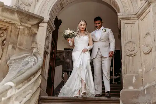 Eine Braut in einem weißen Kleid und ein Bräutigam in einem hellen Anzug schreiten lächelnd die steinernen Stufen unter einem verzierten Torbogen an einem historischen Gebäude hinunter - wunderschön eingefangen von Hochzeitsfotograf Görlitz.