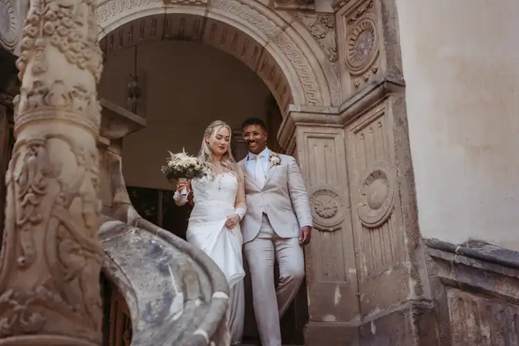 Eine Braut und ein Bräutigam in formeller Hochzeitskleidung gehen lächelnd und händchenhaltend eine Steintreppe auf Hochzeit Gut am See hinunter. Die Braut hält einen Blumenstrauß in der Hand und trägt ein weißes Kleid. Die Szene ist elegant und fröhlich.