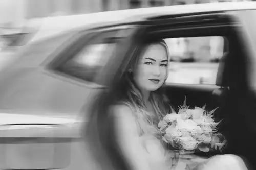Eine Braut im Brautkleid und mit einem Blumenstrauß in der Hand sitzt auf dem Rücksitz eines Autos und blickt aus dem offenen Fenster. Dieses Schwarz-Weiß-Bild von Hochzeitsfotograf Görlitz hat eine weiche, verträumte Unschärfe, die sie einrahmt.