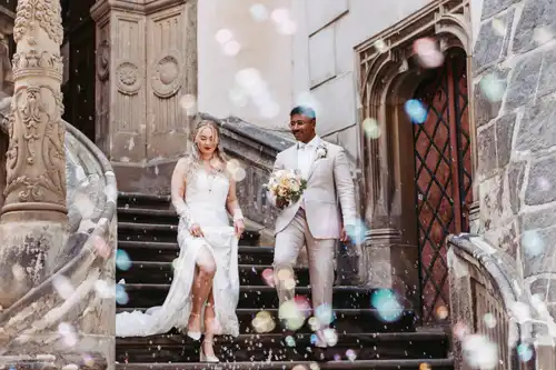 Ein Brautpaar in Hochzeitskleidung schreitet die Steinstufen vor einem historischen Gebäude in Görlitz hinunter, umgeben von buntem Konfetti, das in der Luft schwebt.