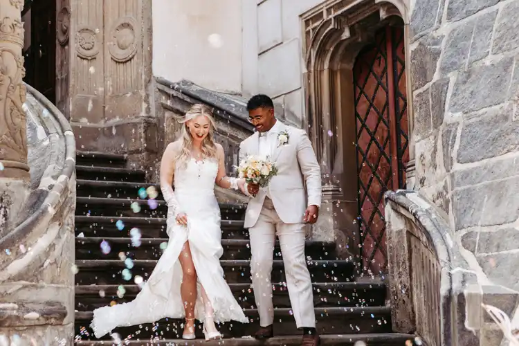 Ein Brautpaar in Hochzeitskleidung schreitet die Steinstufen vor einem historischen Gebäude in Görlitz hinunter, umgeben von buntem Konfetti, das in der Luft schwebt.