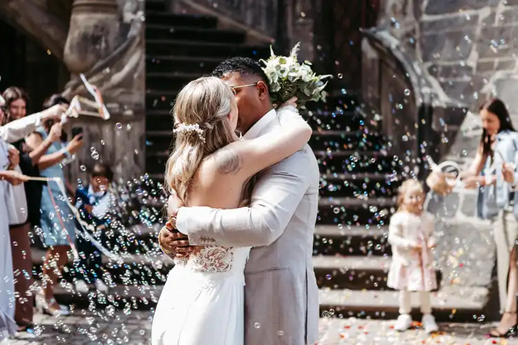 Braut und Bräutigam umarmen und küssen sich bei einer Hochzeit in Görlitz, umgeben von Gästen, die Seifenblasen blasen und ihre Hochzeit vor einer Steintreppe feiern.