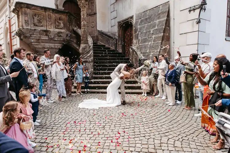 Braut und Bräutigam küssen sich leidenschaftlich auf einer Kopfsteinpflasterstraße vor einem historischen Gebäude in Görlitz, während die Gäste sie mit Blumenblättern überschütten - wunderschön eingefangen von Hochzeitsfotograf Görlitz inmitten einer fröhlichen Feier.