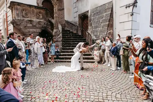 Braut und Bräutigam küssen sich leidenschaftlich auf einer Kopfsteinpflasterstraße vor einem historischen Gebäude in Görlitz, während die Gäste sie mit Blumenblättern überschütten - wunderschön eingefangen von Hochzeitsfotograf Görlitz inmitten einer fröhlichen Feier.