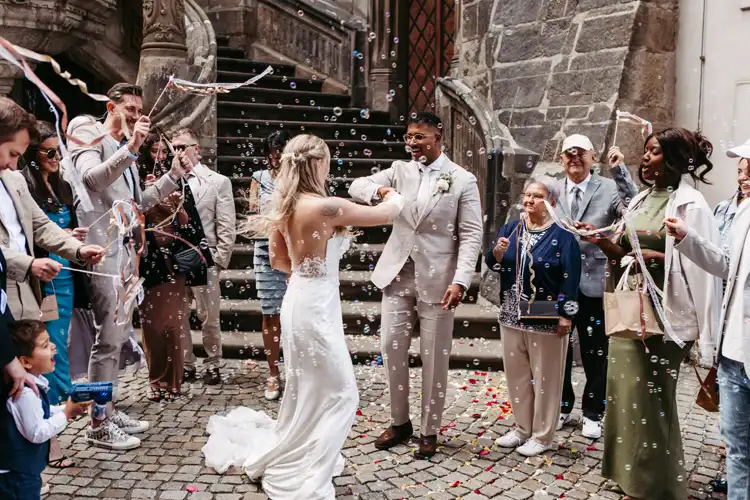 Ein frisch verheiratetes Paar umarmt sich und lächelt bei seiner Hochzeit in Görlitz umgeben von jubelnden Gästen, die in einem mit Steinen gepflasterten Innenhof Seifenblasen blasen und mit Bändern winken. Alle sehen fröhlich und festlich aus.