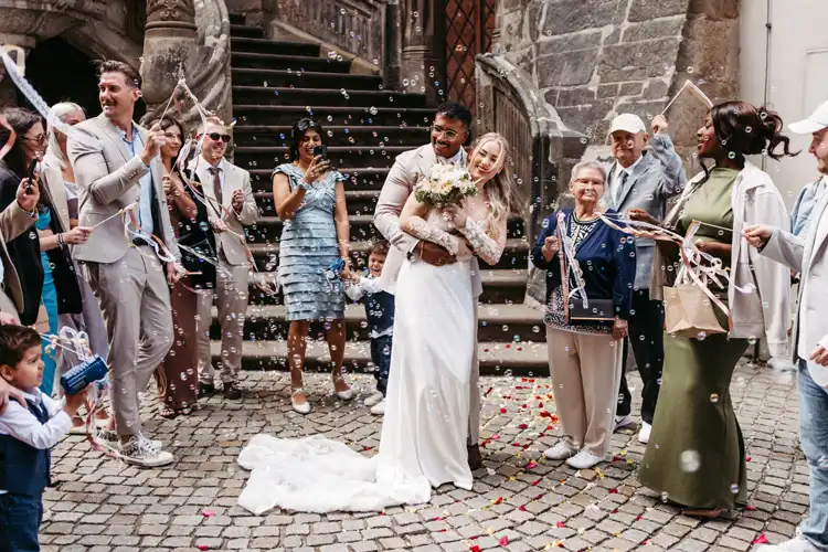 Ein frisch verheiratetes Paar umarmt sich und lächelt bei seiner Hochzeit in Görlitz umgeben von jubelnden Gästen, die in einem mit Steinen gepflasterten Innenhof Seifenblasen blasen und mit Bändern winken. Alle sehen fröhlich und festlich aus.