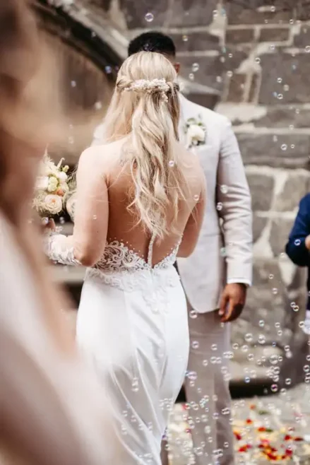 Eine Braut mit langen blonden Haaren in einem weißen, rückenfreien Spitzenkleid steht ihrem Bräutigam in einem hellen Anzug gegenüber. Im Freien auf Hochzeit in Görlitz sind sie von einer Steinmauer umgeben und werden von Seifenblasen und Blumenblättern umringt.