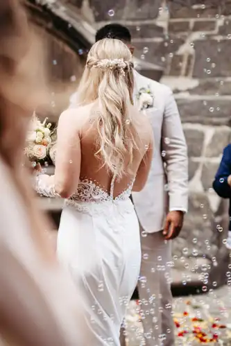 Eine Braut mit langen blonden Haaren in einem weißen, rückenfreien Spitzenkleid steht ihrem Bräutigam in einem hellen Anzug gegenüber. Im Freien auf Hochzeit in Görlitz sind sie von einer Steinmauer umgeben und werden von Seifenblasen und Blumenblättern umringt.