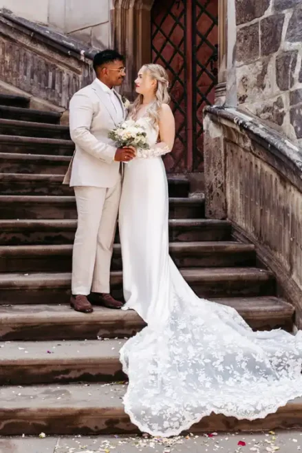 Eine Braut in einem langen weißen Kleid mit Spitzenschleppe und ein Bräutigam in einem hellen Anzug stehen auf den Steinstufen vor einem Gebäude, schauen sich an und halten einen Blumenstrauß in der Hand - wunderschön eingefangen von Hochzeitsfotograf Görlitz.