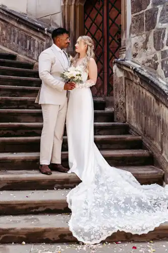 Eine Braut in einem langen weißen Kleid mit Spitzenschleppe und ein Bräutigam in einem hellen Anzug stehen auf den Steinstufen vor einem Gebäude, schauen sich an und halten einen Blumenstrauß in der Hand - wunderschön eingefangen von Hochzeitsfotograf Görlitz.