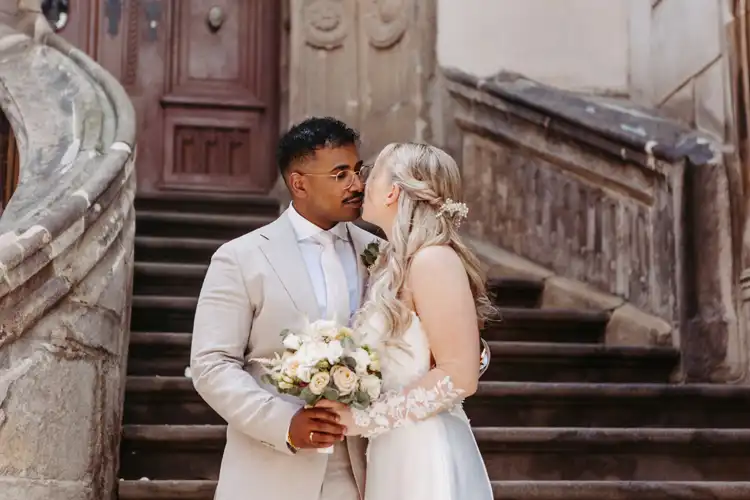 Eine Braut und ein Bräutigam stehen dicht beieinander auf den alten Steinstufen der Hochzeit Gut am See und blicken sich in die Augen. Der Bräutigam trägt einen hellen Anzug und eine Brille und hält einen Blumenstrauß in der Hand. Die Braut hat langes blondes Haar und trägt ein weißes Kleid.