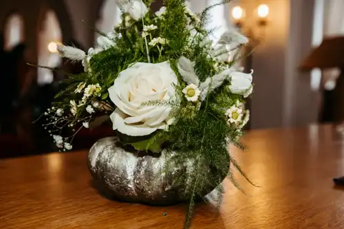Eine silberne Vase hält ein Blumenarrangement mit einer großen weißen Rose, kleinen weißen Blumen und grünem Laub. Es steht auf einem Holztisch im Innenbereich des Standesamtes Görlitz, der in sanftes Licht getaucht ist - perfekt für einen Hochzeitsfotografen Görlitz, um es einzufangen.