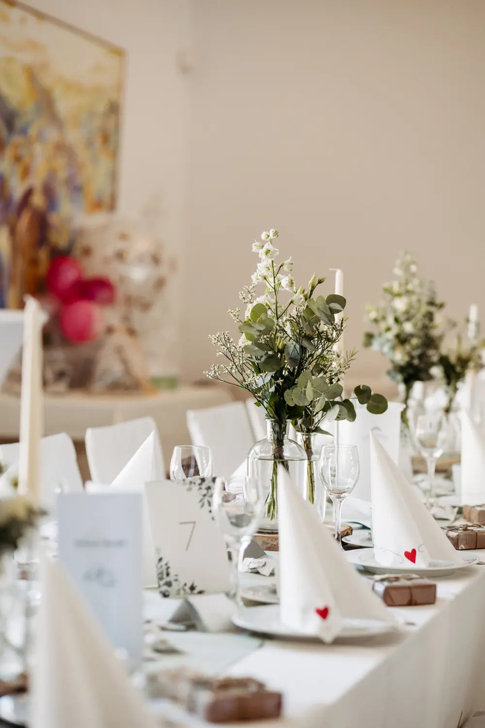 Eine lange Banketttafel ist elegant mit weißen, zu Dreiecken gefalteten Servietten, Gläsern, Tellern, grünen Blumenkränzen und nummerierten Tischkarten gedeckt. Kleine Geschenkboxen und dekorative Akzente schaffen eine festliche Atmosphäre.